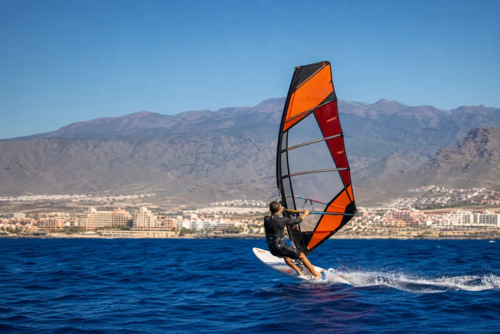 Realistyczne zdjęcie ukazujące windsurfera na desce z pomarańczowo-czerwonym żaglem na tle wybrzeża Costa Adeje i gór południowej Teneryfy. Dominują żywe, naturalne kolory — głęboki błękit morza, kontrastujący pomarańcz żagla i stonowane brązy gór; oświetlenie dzienne i naturalna perspektywa podkreślają fotograficzny styl. Kompozycja z zawodnikiem przesuniętym na prawo (zasada trzech części) czyni obraz idealnym jako zdjęcie nagłówkowe do artykułu, poradnika lub przewodnika o windsurfingu i warunkach pogodowych.